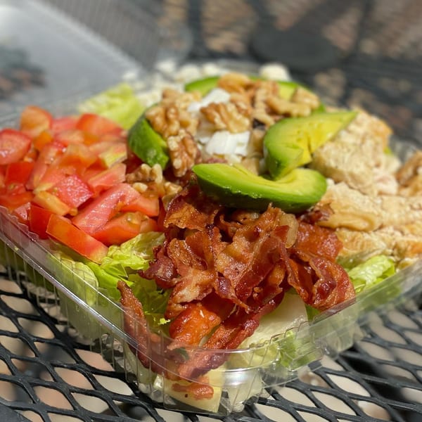 A Sandwich Club salad with lettuce, tomatoes, bacon, avocado, walnuts, and chicken in a plastic container.